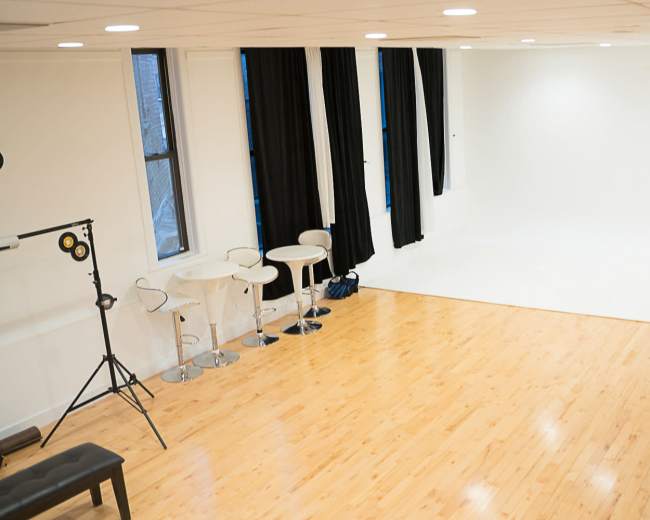 The image shows a spacious, empty studio with a polished wooden floor, white walls, large windows, and black curtains, along with minimal furnishings including a seating area and a small table.