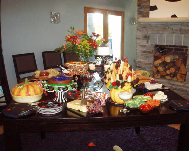 A large dining table is set with various food items, including cakes, fruits, and a decorative centerpiece of flowers, with a stone fireplace in the background.
