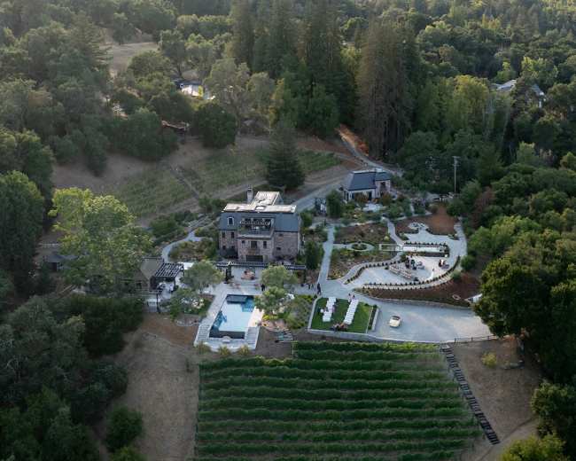 An aerial view shows a vineyard estate with a modern house, swimming pool, and surrounding landscaping nestled among rolling hills and trees.