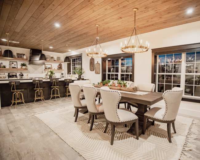 The image shows a modern dining room with a rectangular wooden table surrounded by upholstered chairs, positioned next to a kitchen area with bar stools and large windows.