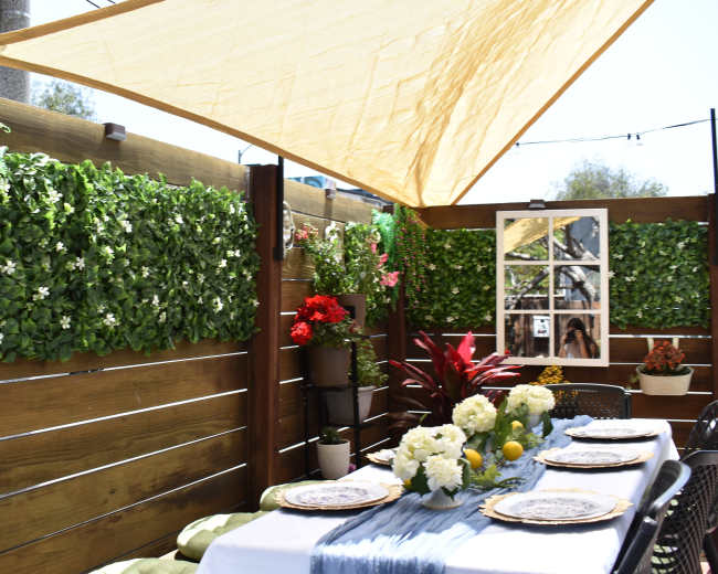 A sunny outdoor patio features a table set with plates and flowers, surrounded by green foliage and decorative plants against wooden walls.