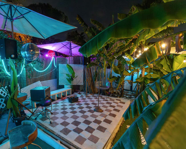 The image shows a colorful outdoor entertainment area featuring a checkered floor, a stage with a microphone, and illuminated decorations among tropical plants.