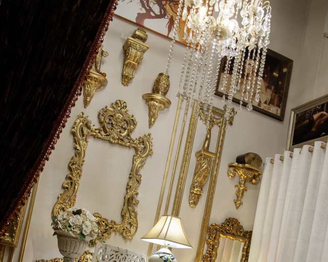 The image features an ornate white desk adorned with intricate carvings, set against a backdrop of gold-framed mirrors and decorative wall accents in a lavishly styled room.