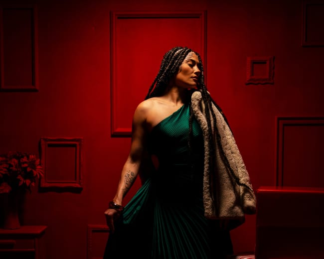A woman wearing a one-shoulder green dress and a fur coat stands among wrapped Christmas gifts against a red backdrop.