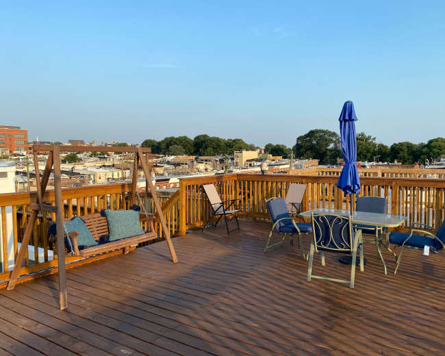 A rooftop deck with a wooden swing, several chairs, a table, and an umbrella, overlooking a cityscape.