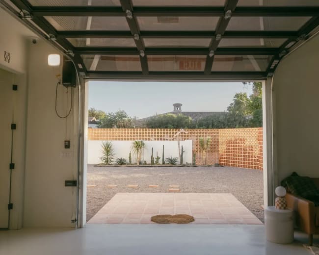 A wide garage door opens to a gravel yard with landscaped plants and a wall in the background.