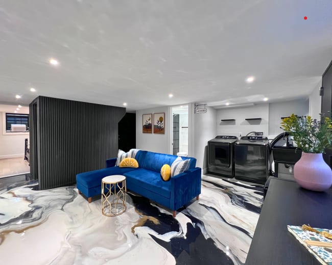 The image shows a modern basement living area featuring a blue sectional sofa, a small round table, and laundry machines in the background against a polished marble floor.