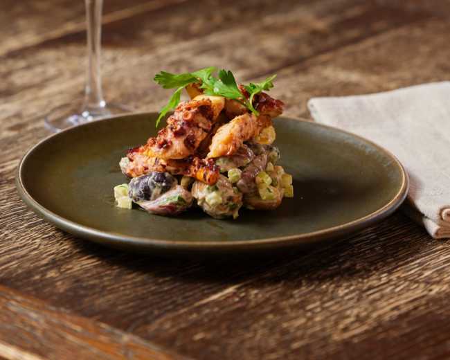 A plate of fried chicken is served atop a creamy salad with vegetables, accompanied by a glass of white wine on a wooden table.