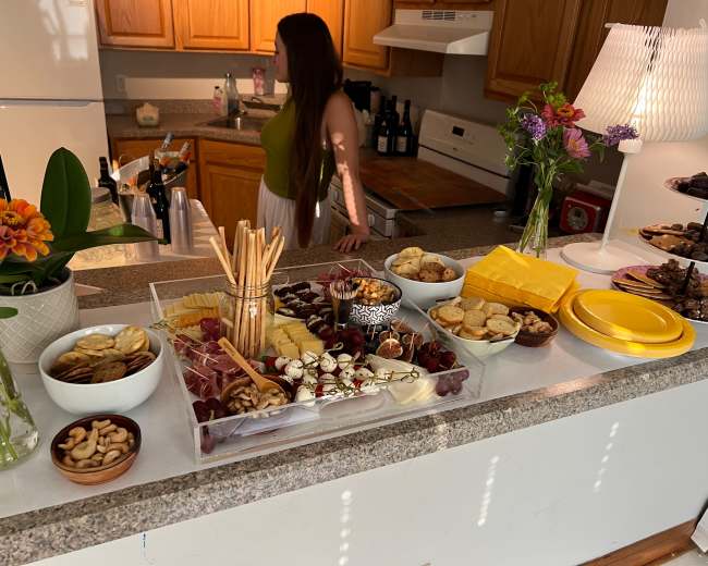A table is set with an array of snacks including fruits, cheeses, crackers, and cookies, while a person stands in the background near the kitchen.