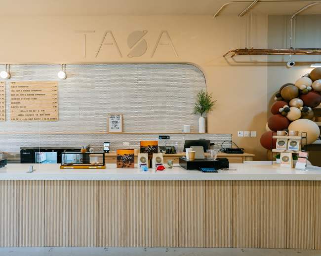 A modern coffee shop counter with a light-colored wooden facade, various menu boards, and decorative elements including a display of coffee products and a plant.