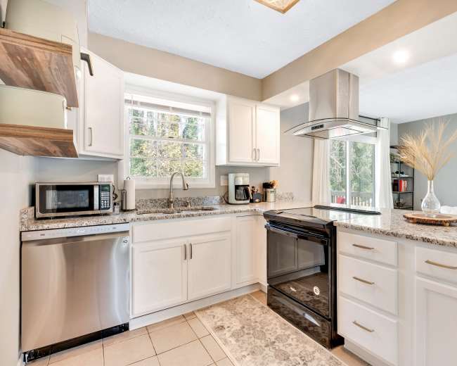 The image shows a modern kitchen with white cabinets, stainless steel appliances, and a window overlooking a garden area.