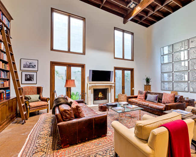 The image shows a spacious living room with high ceilings, a fireplace, a large bookshelf, and various seating arrangements, including leather couches and armchairs.