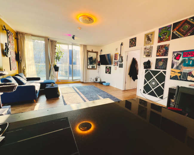 The image shows a brightly lit living room with a blue couch, a large window, a flat-screen TV, and colorful wall art.