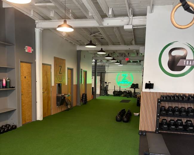The image shows a spacious gym interior with artificial turf flooring, shelves for weights, and kettlebells organized against the wall, along with a workout bench and exercise equipment in the background.