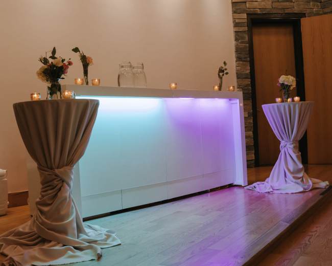 A sleek, illuminated bar is flanked by two draped tables adorned with flowers and candles in a well-lit indoor setting.