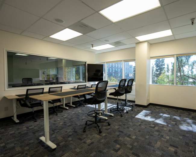 The image shows a modern office space with a conference table, several chairs, large windows, and a view of trees outside.