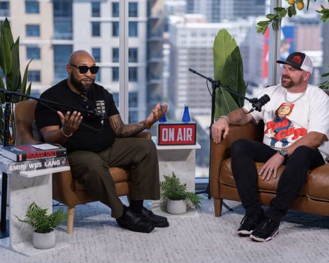 Two men are seated in a modern studio setting with large windows revealing a cityscape, engaged in conversation while surrounded by plants and decorative elements.