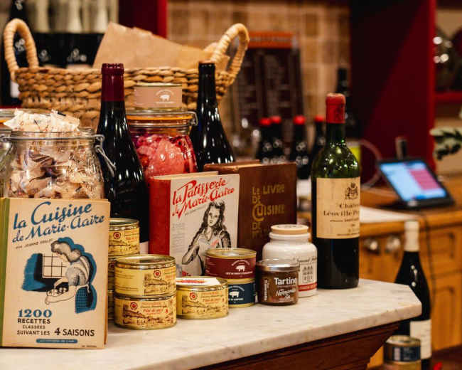The image shows a countertop filled with various food items, including jars, bottles of wine, and canned products, all arranged alongside a decorative basket.