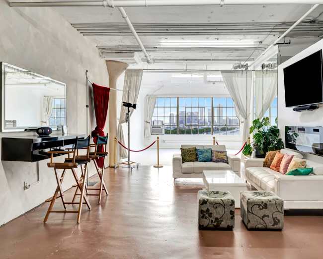 The image shows a spacious, modern living area with large windows, a white couch adorned with colorful pillows, a mirrored vanity, and a flat-screen television mounted on the wall.