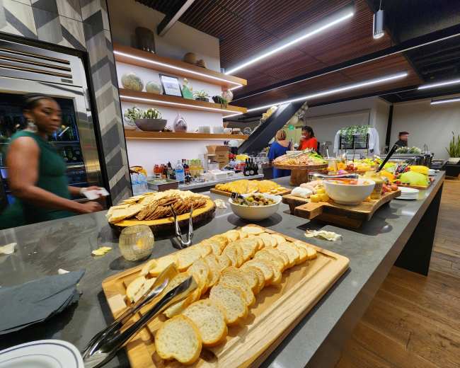 A buffet table displays a variety of foods, including bread, salads, fruits, and appetizers, while individuals prepare and serve the items in a modern kitchen setting.