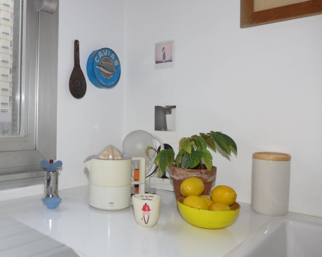 A kitchen counter features a coffee maker, a bowl of lemons, and various kitchen utensils near a window overlooking a building.