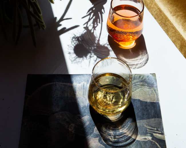 Two glasses of drinks are placed on a table beside a vase with flowers, casting shadows on the surface, illuminated by sunlight.
