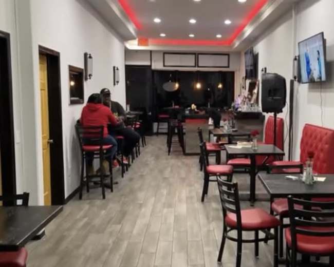 A narrow restaurant interior with several tables and chairs, and patrons sitting at the bar along one side.
