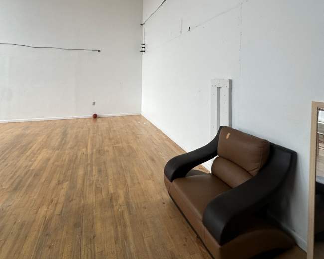 An empty room with wooden flooring features a brown and black sofa against a white wall.