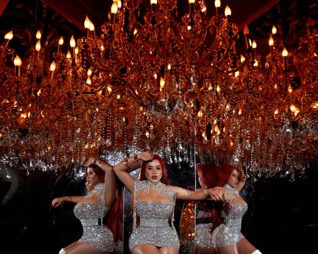 A woman in a sparkling silver dress and white knee-high boots poses confidently in front of a large ornate chandelier, with her reflection visible in multiple mirrors.