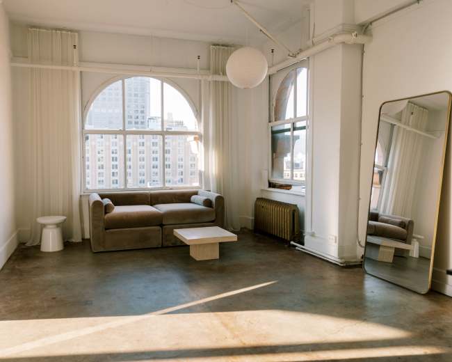 The image shows a spacious, well-lit living area featuring a beige sofa, a small white table, a large mirror, and a window with city views.