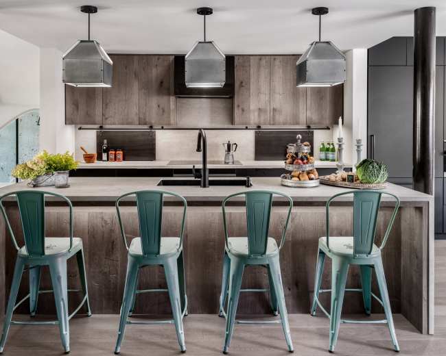 A modern kitchen features a large island with four metal stools, dark wood cabinetry, and pendant lights above.