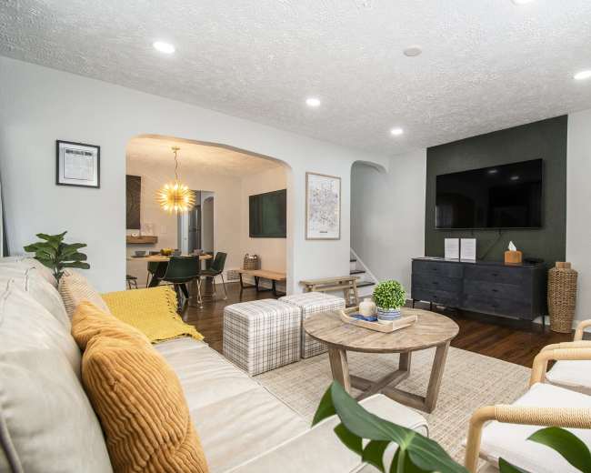 The living room features a light-colored sofa with yellow and orange throw pillows, a wooden coffee table, and a wall-mounted television, with an open archway leading to a dining area.