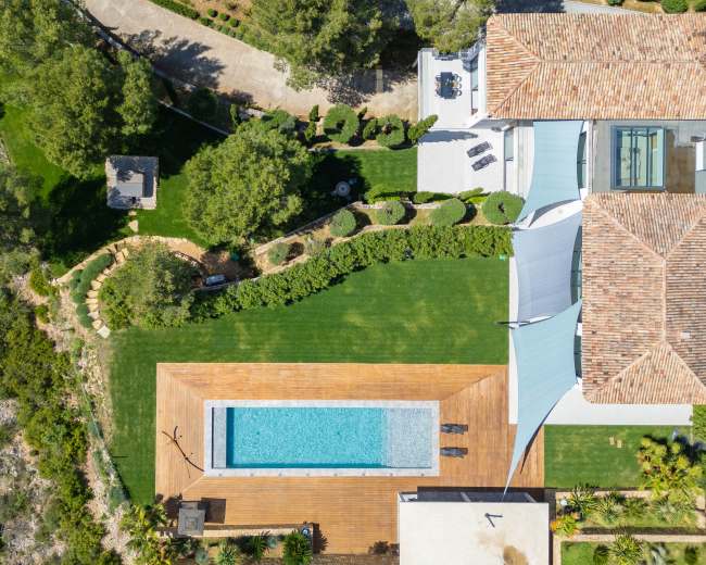 The image shows an aerial view of a backyard featuring a swimming pool surrounded by wooden decking, landscaped gardens, and a house with a tiled roof.