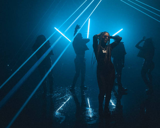 A group of silhouetted dancers illuminated by bright blue laser beams in a dimly lit space.
