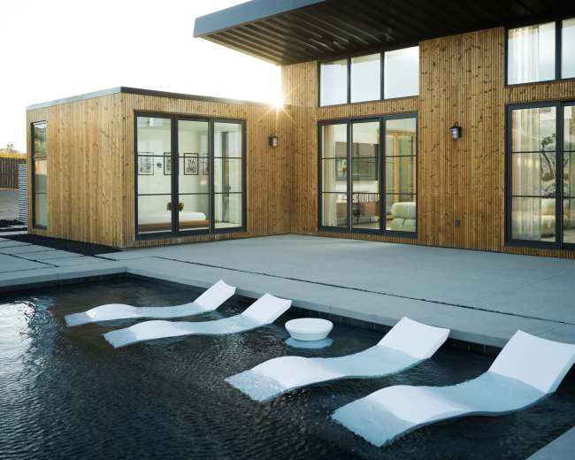 The image shows a modern home with a wooden exterior, featuring a pool with white lounge chairs partially submerged in the water.