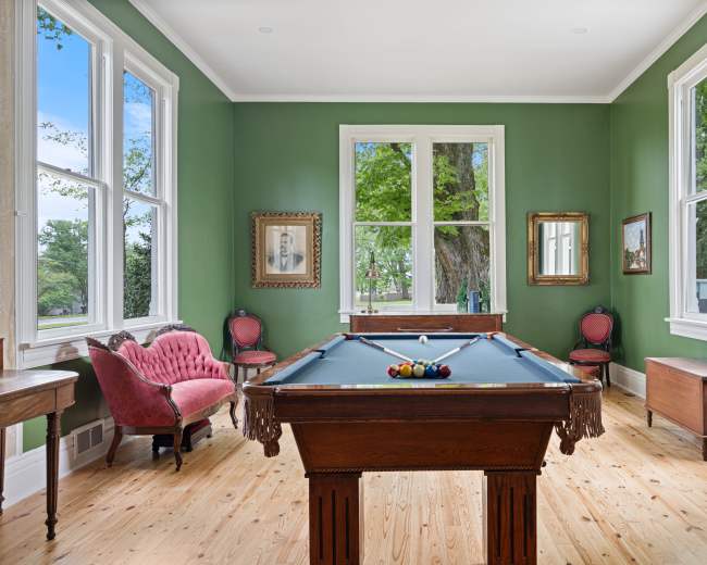 The image shows a billiard room with a green wall, a pool table in the center, and large windows that overlook a garden.
