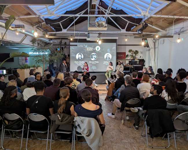 A diverse audience sits in a well-lit room while listening to speakers at a panel event on a stage.