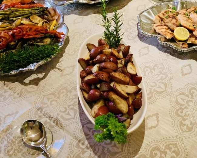 The table is set with platters of grilled meats and roasted vegetables, accompanied by a bowl of seasoned red potatoes garnished with parsley and a sprig of rosemary.