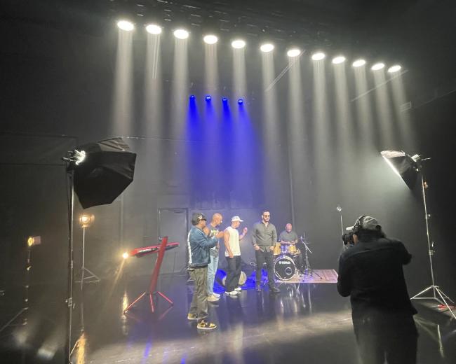 A band performs in a dimly lit studio with bright lights overhead and a photographer capturing the scene.