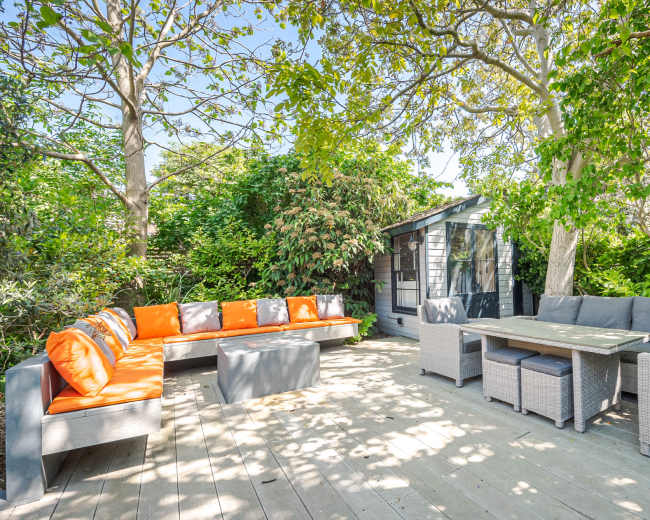 A patio area features a seating arrangement with orange and gray cushions, surrounded by lush greenery and a small shed in the background.