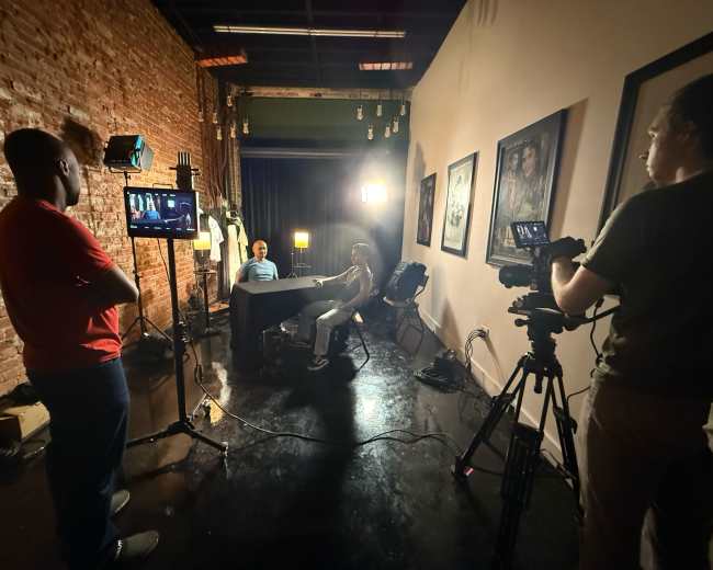 A film crew sets up a scene in a brick-walled studio, featuring two seated actors at a table under bright lights, while crew members operate cameras and monitor equipment.