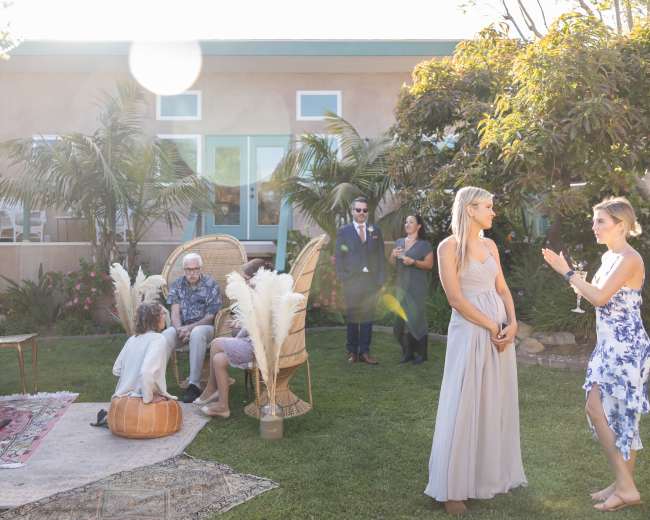 A group of people mingles outdoors in a garden setting, with decorative furniture and greenery surrounding them.
