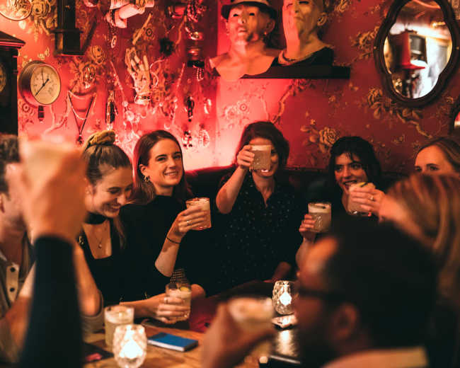 A group of people is raising their drinks in a dimly lit bar decorated with vintage clocks and masks.