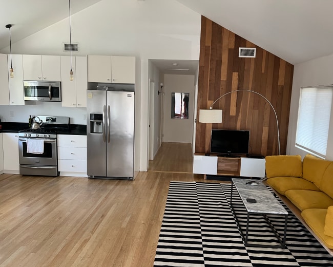 The image shows a modern open-concept living area with a kitchen, a yellow sofa, a striped rug, and a wood-paneled accent wall.