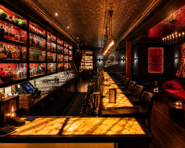 The image features an elegantly designed bar interior with illuminated shelves displaying an array of spirits and a long, glowing bar counter.