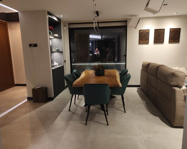 The image shows a modern dining area featuring a wooden table surrounded by six teal chairs, with a cozy couch and decorative pieces on the walls.