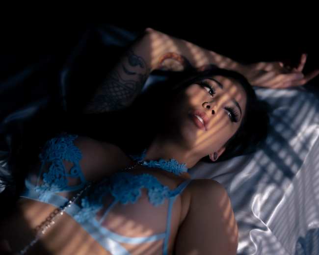 A person lies on a satin surface with their arms stretched above their head, partially illuminated by shadowy patterns.