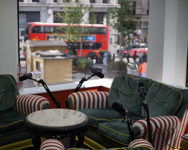 The image shows two striped armchairs facing a round table, with a view of a busy street and a red double-decker bus visible through a large window.