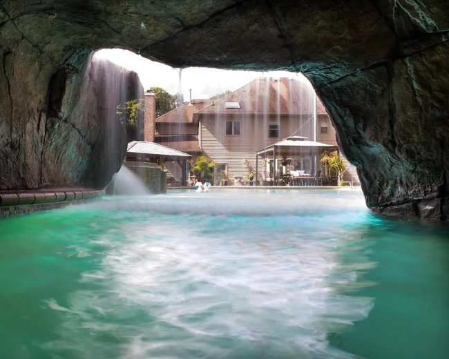 A view through a rocky cave entrance reveals a swimming pool surrounded by a house and outdoor patio area.