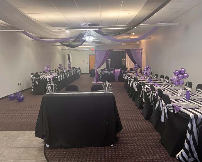 A decorated event space with black and purple table settings, balloons, and draped fabric overhead.